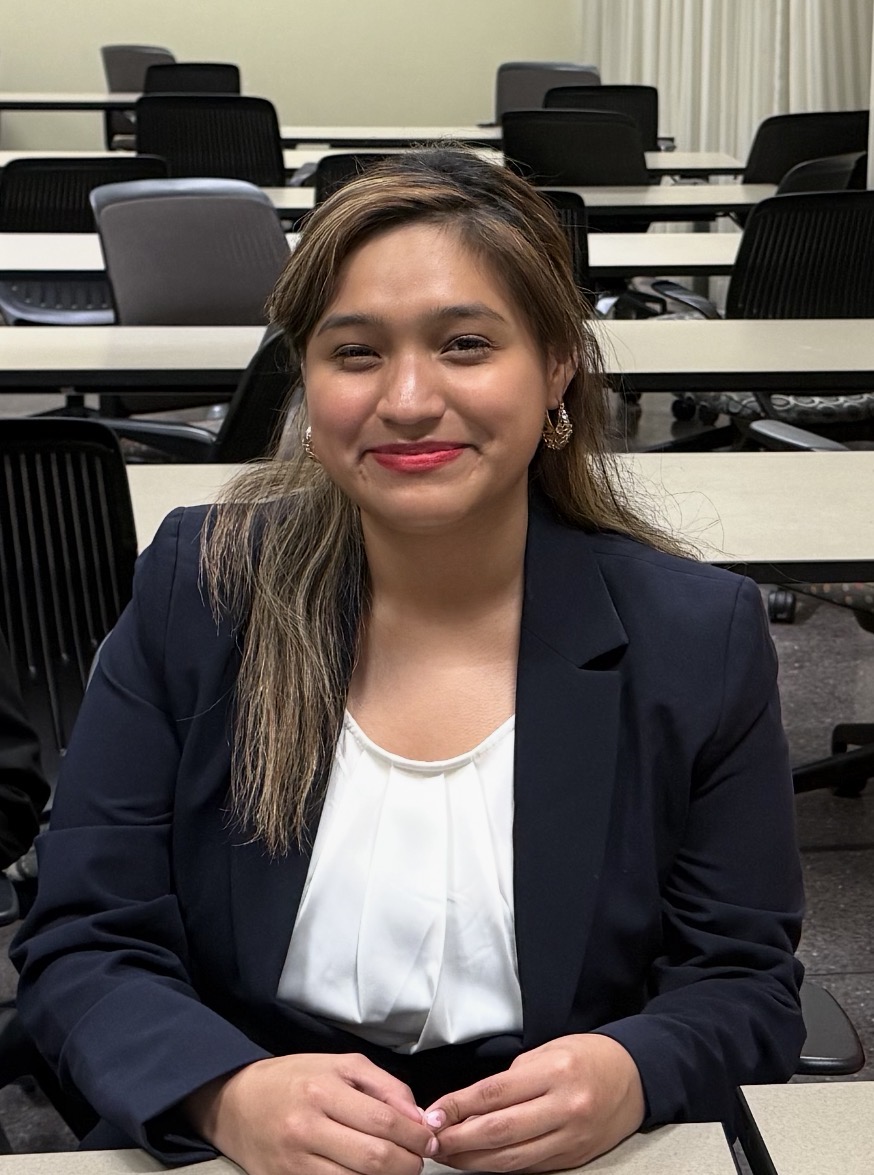 Andrea Hernandez, CEO and Founder of AccessWave Technologies, sitting in a conference room. She has medium-long straight dark brown hair with blonde highlights framing her face, dark brown eyes, and is smiling gently while wearing pink-toned lipstick and small gold dangling earrings. She is dressed in a navy blue blazer over a white pleated blouse, with her hands clasped together resting on a table in front of her. The background shows rows of empty black rolling chairs and beige folding tables of a conference room.