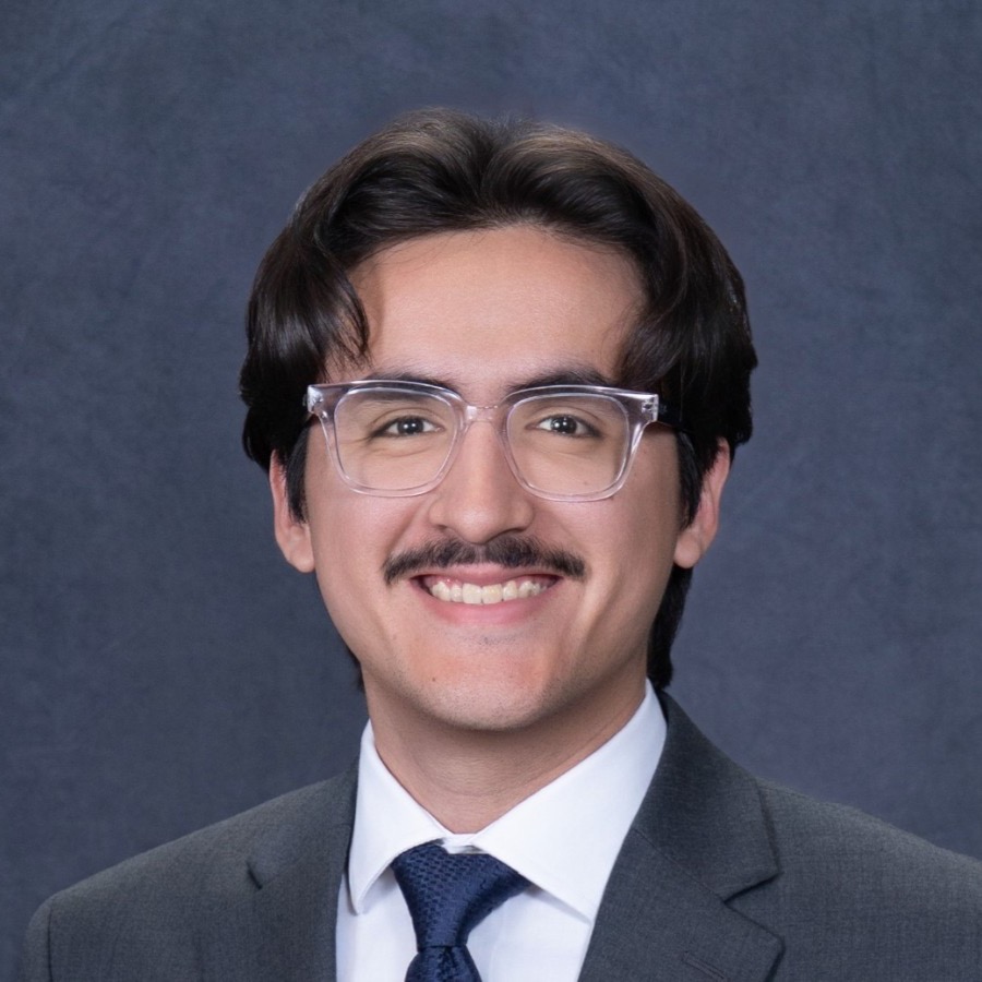 Professional headshot of Andres Corrales, Senior Engineer. He has medium-length black hair styled in a middle part, a black mustache, and wears clear-framed glasses, looking directly at the camera. He is dressed in a gray suit jacket over a white collared shirt with a navy tie. The background is a plain studio backdrop.