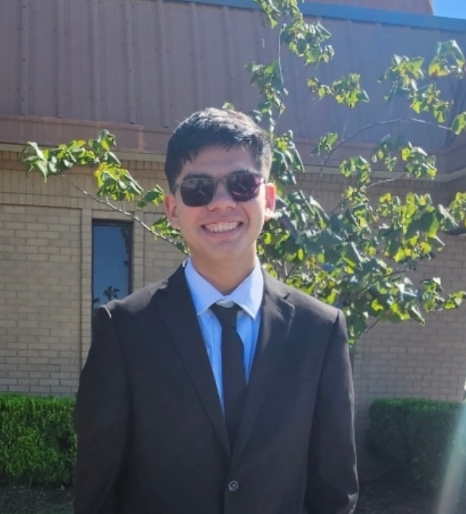 Ian Flores, Founding Engineer, outdoors with short dark brown hair, smiling broadly with his teeth showing. He is wearing dark sunglasses, a white collared dress shirt, a black necktie, and a black suit jacket, standing facing the camera with one arm relaxed at his side. Behind him is a tan brick building and a green leafy tree branch extending across the upper right portion of the image.