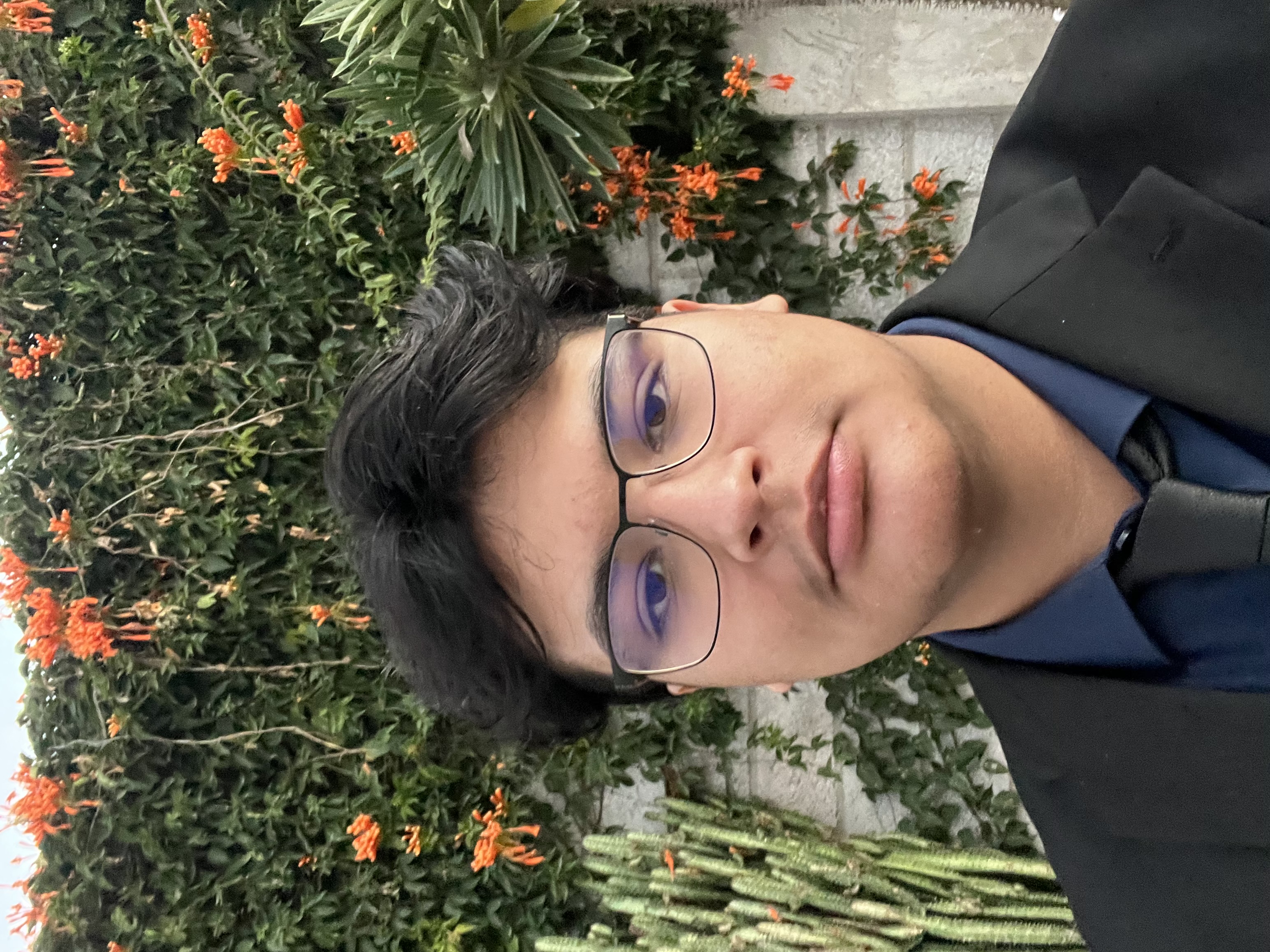 Jesus Cervantes, CTO and co-founder, standing outdoors and facing the camera with a neutral expression. He has short dark brown hair, dark brown eyes, and wears rectangular black glasses. He is dressed in a black suit jacket over a dark navy button-down shirt with a black tie. Behind him is a light stone wall covered with green climbing vines and bright orange trumpet-shaped flowers, with a tall cactus to the left and a large green succulent plant to the right.