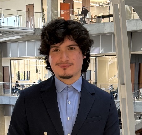 Joshua Chavarría, founding engineer, in the Engineering Education and Research Center building at UT Austin. He has dark curly, wavy, shoulder-length hair, hazel eyes, and light facial hair along his jaw and upper lip, smiling and looking directly at the camera. He is wearing a navy blue blazer over a light blue button-up collared shirt. The background shows the building’s modern multi-story interior with glass and large windows letting in natural light.