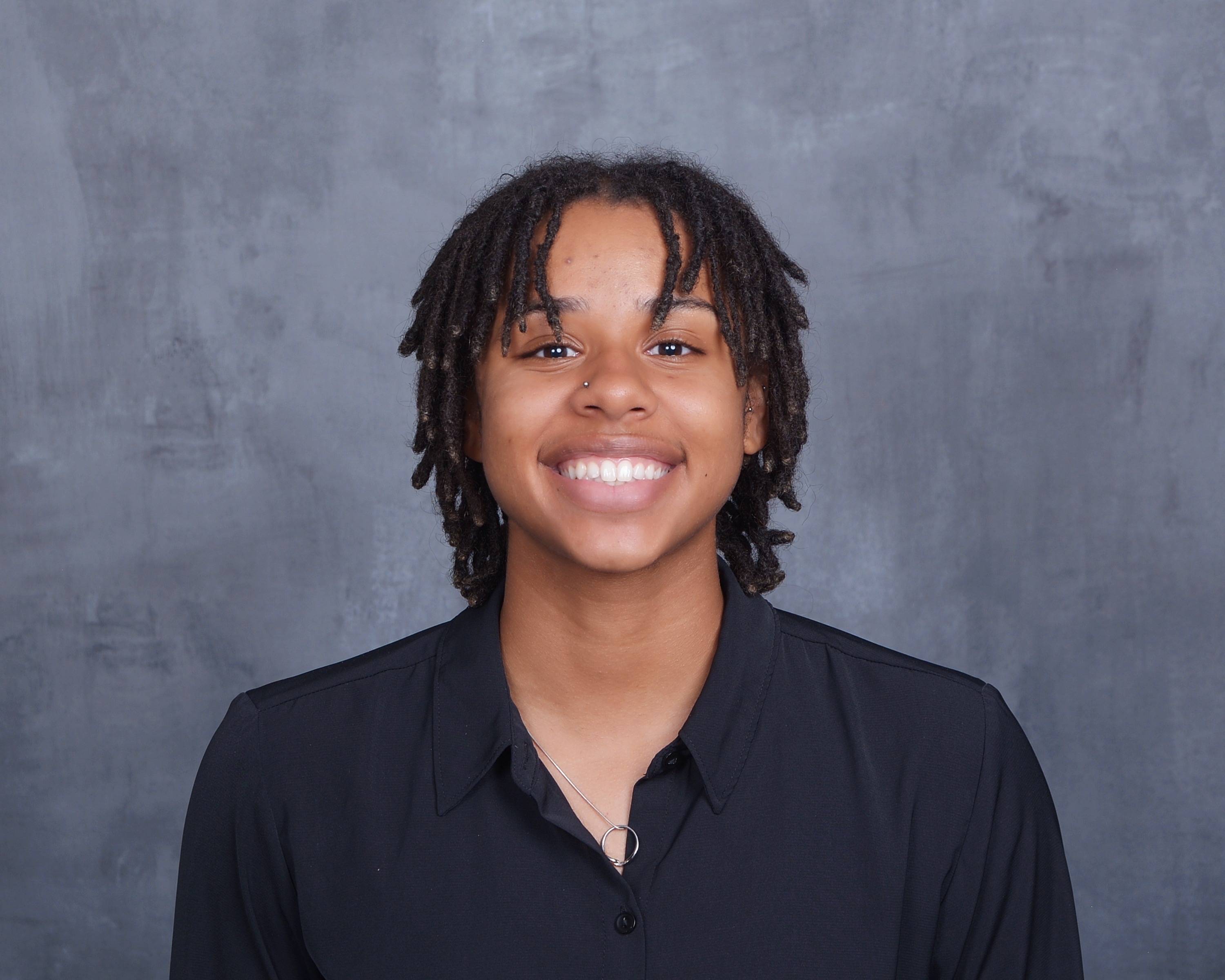 Headshot of Olyvia Witham, Founding Engineer, smiling warmly with her teeth showing. She has shoulder-length, dark brown loc’d (dreadlocked) hair parted to frame her face, dark brown eyes, a small nose ring on her left nostril, and small stud earrings. She is wearing a solid black button-up collared shirt with a thin silver chain necklace. The background is a plain, mottled gray studio backdrop.